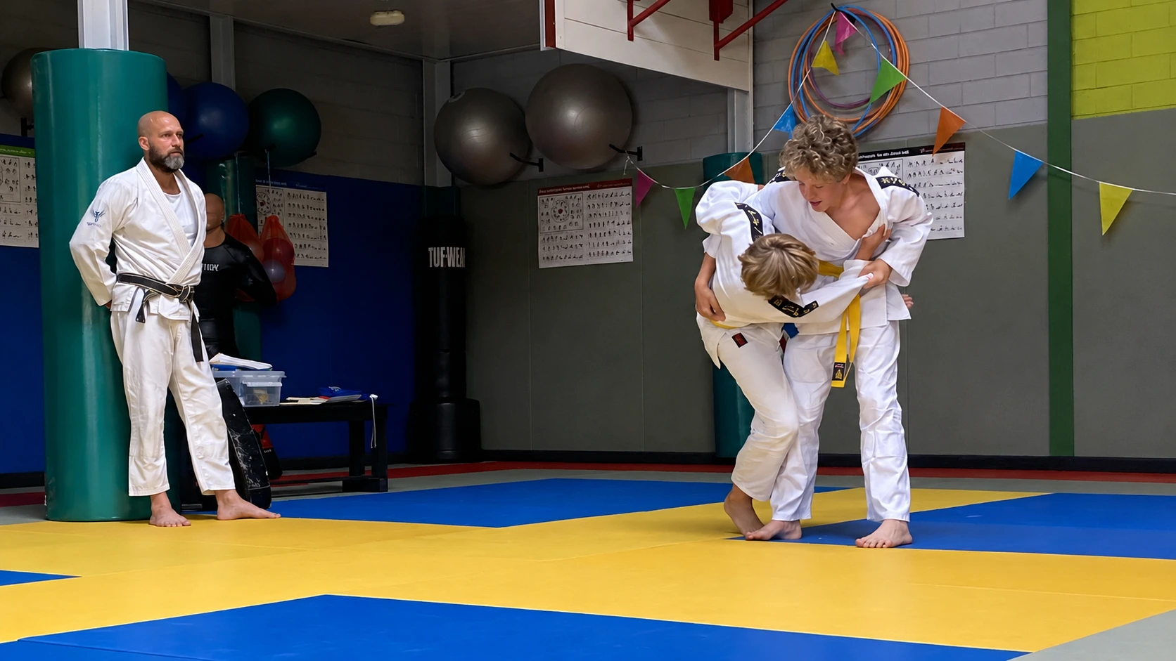 Judo voor kinderen in Hardinxveld-Giessendam Twee jeugdjudoka's oefenen een worp terwijl de trainer meekijkt.